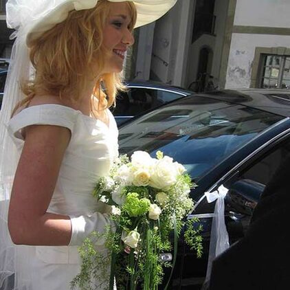 Braut mit Hut und einem weiß-grünen Brautstrauß neben dem Hochzeitsauto