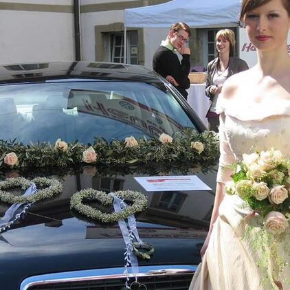 Braut vor einem Auto mit einem Rosenherz auf der Motorhaube